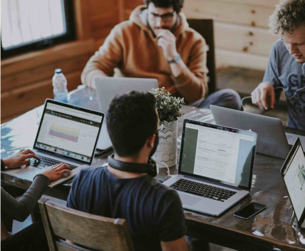 Group of people engaged in collaborative work at a wooden table with laptops, smartphones, and a calculator, indicating a casual business meeting or co-working environment